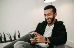 Indian man playing at an online casino on his smartphone. Laughing, smiling, wearing white shirt, black hoodie.