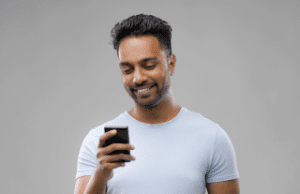 Indian man playing at an online casino on his smartphone. Smiling, wearing light blue shirt.
